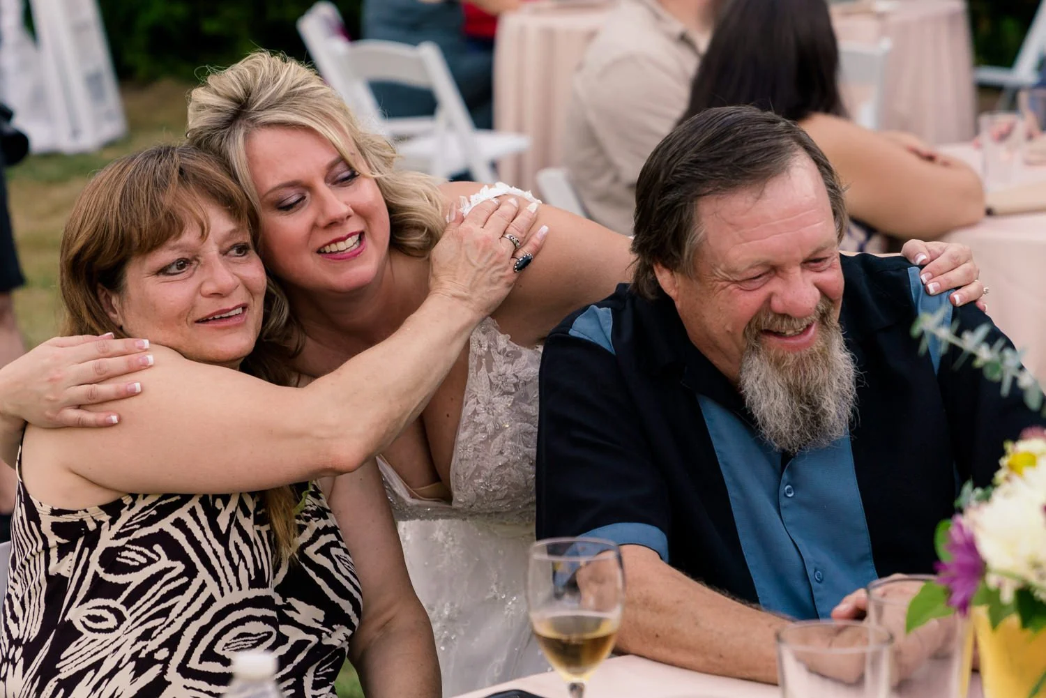 Bride hugging her parents during an emotional moment at a backyard wedding reception in Vancouver Washington, candid family photo with wine glasses and floral centerpieces on the table.
