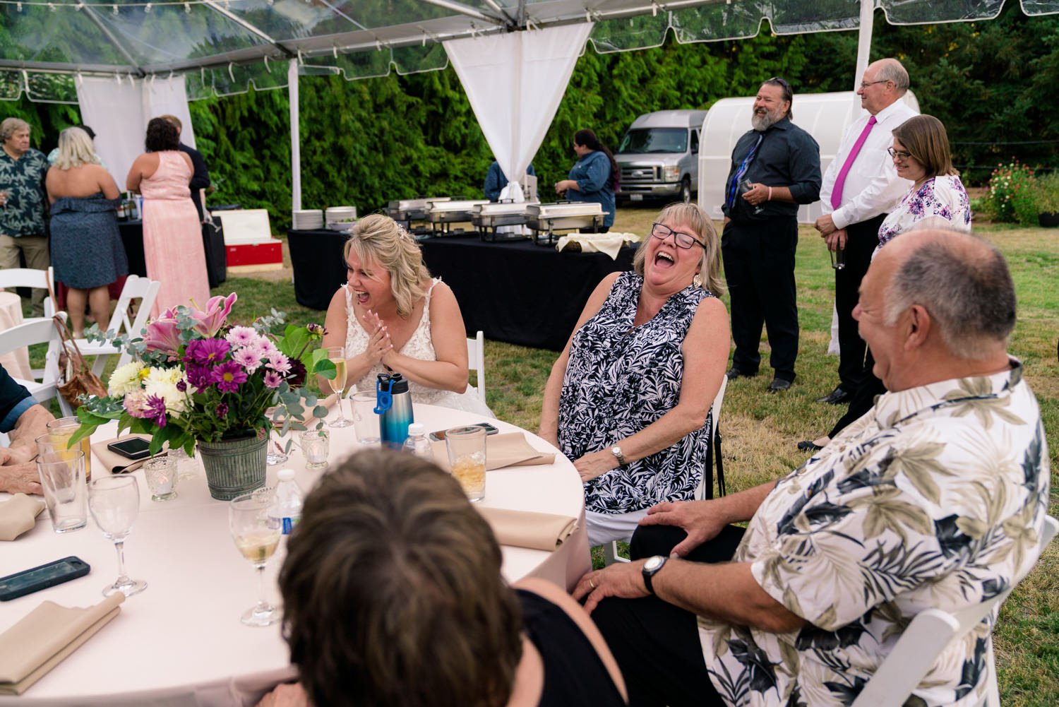 Guests laughing together under a clear tent at a Vancouver WA backyard wedding reception, candid moment with round tables, floral centerpiece, and relaxed outdoor celebration vibe.