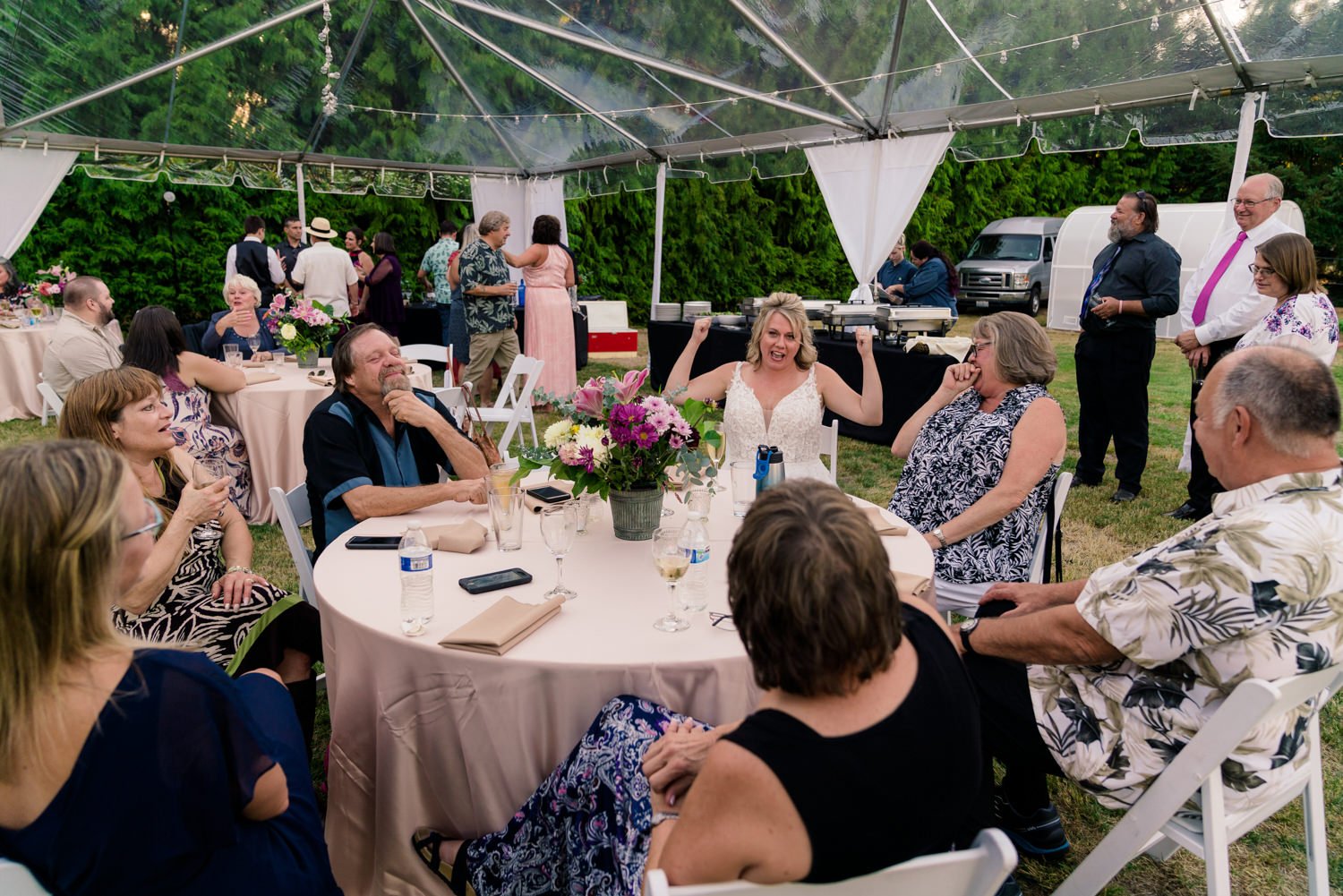 Bride cheering with arms raised while guests sit around round tables under a clear tent at a Vancouver Washington backyard wedding reception filled with laughter and celebration.