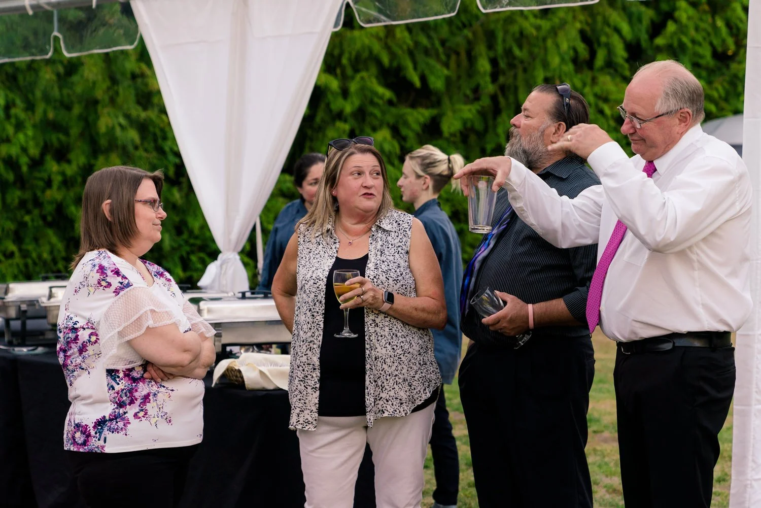 Wedding guests chatting and holding drinks near the buffet line under a tent at a Vancouver WA backyard wedding reception with evergreen trees in the background.