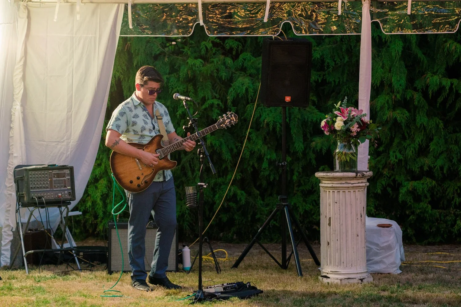 Live musician playing guitar and singing under a reception tent at a Vancouver Washington backyard wedding, outdoor entertainment with speakers and greenery behind the stage area.