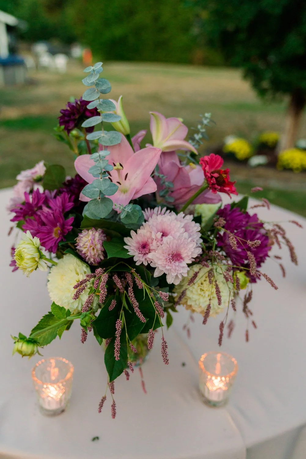 Colorful wedding floral centerpiece with pink lilies, purple mums, and greenery on a round reception table at a Vancouver WA backyard wedding with candlelight details.