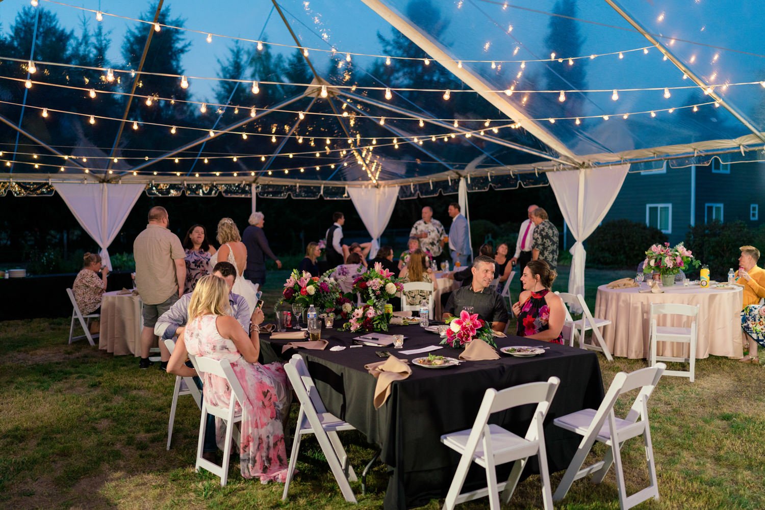 Guests gathered under a clear tent with glowing string lights at a backyard wedding in Vancouver Washington, evening reception with round tables, florals, and relaxed outdoor celebration.