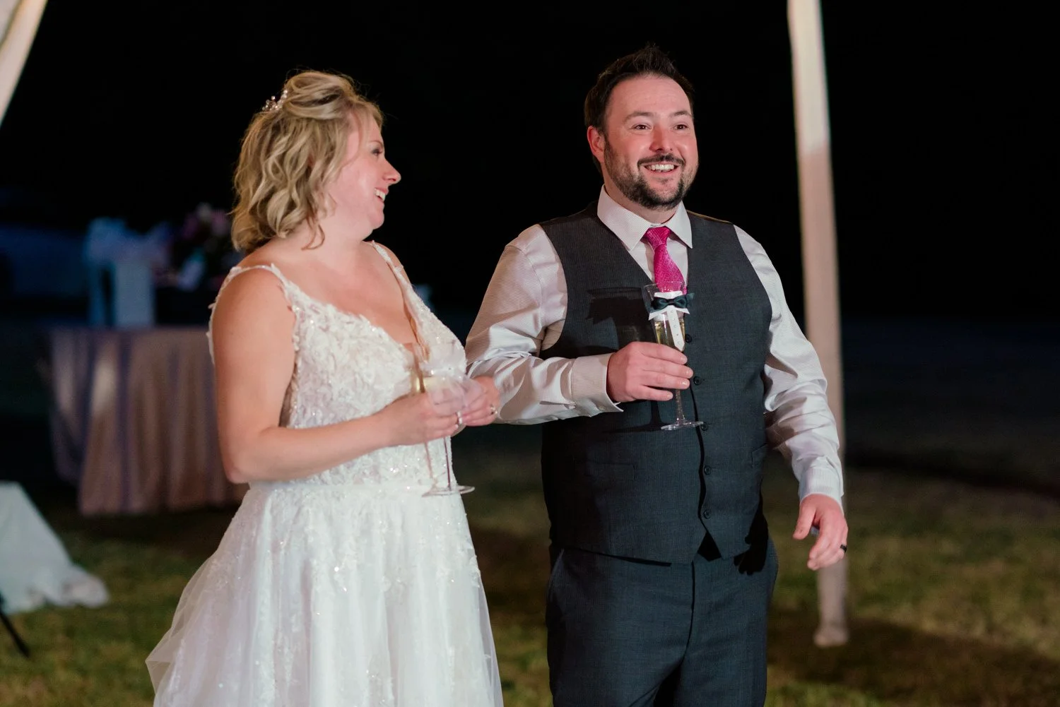 Bride and groom smiling with champagne glasses during their Vancouver WA backyard wedding reception at night, candid moment under tent lighting and open sky.
