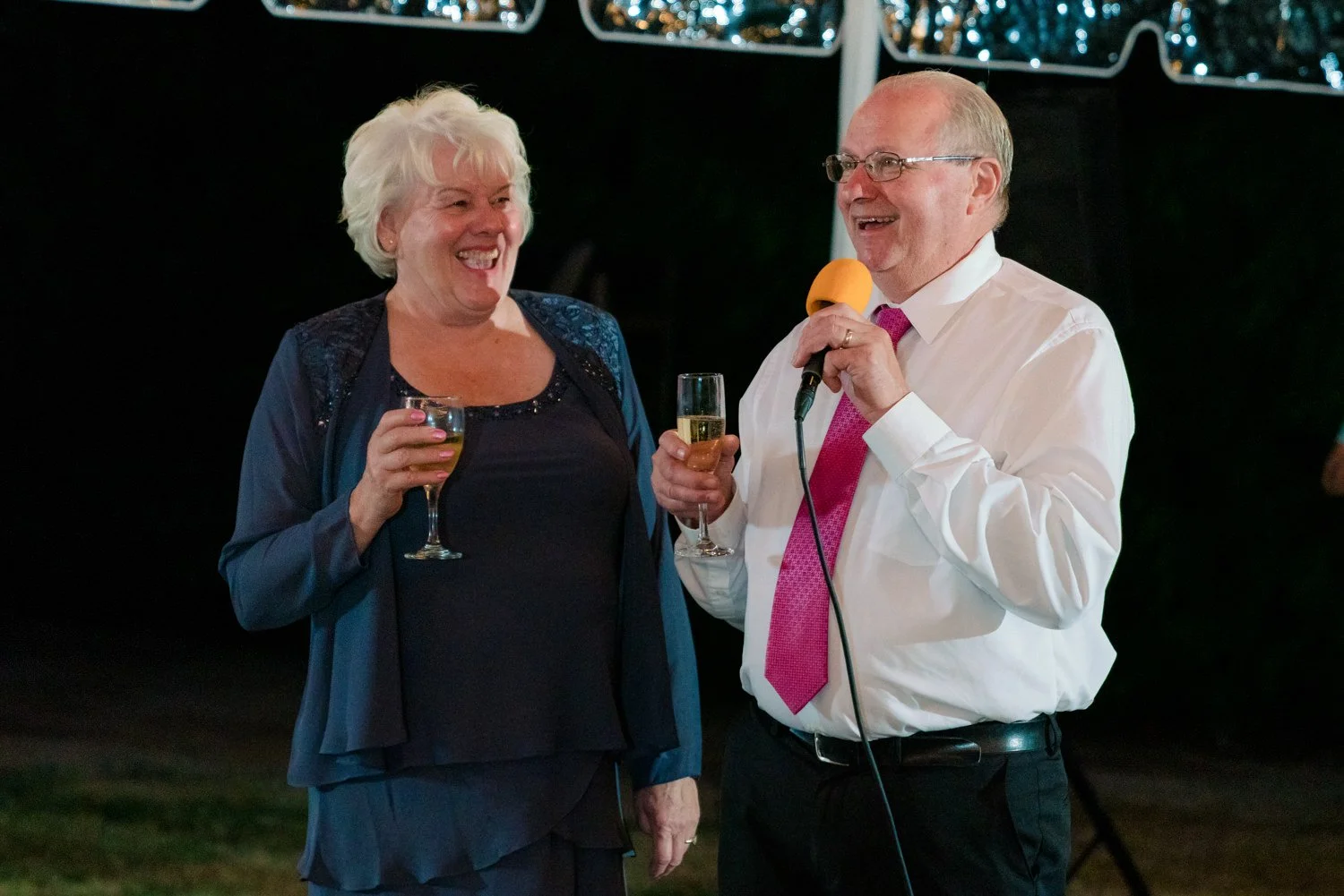 Father of the bride giving a heartfelt speech with microphone while mother smiles beside him at a Vancouver Washington backyard wedding reception under twinkle lights.