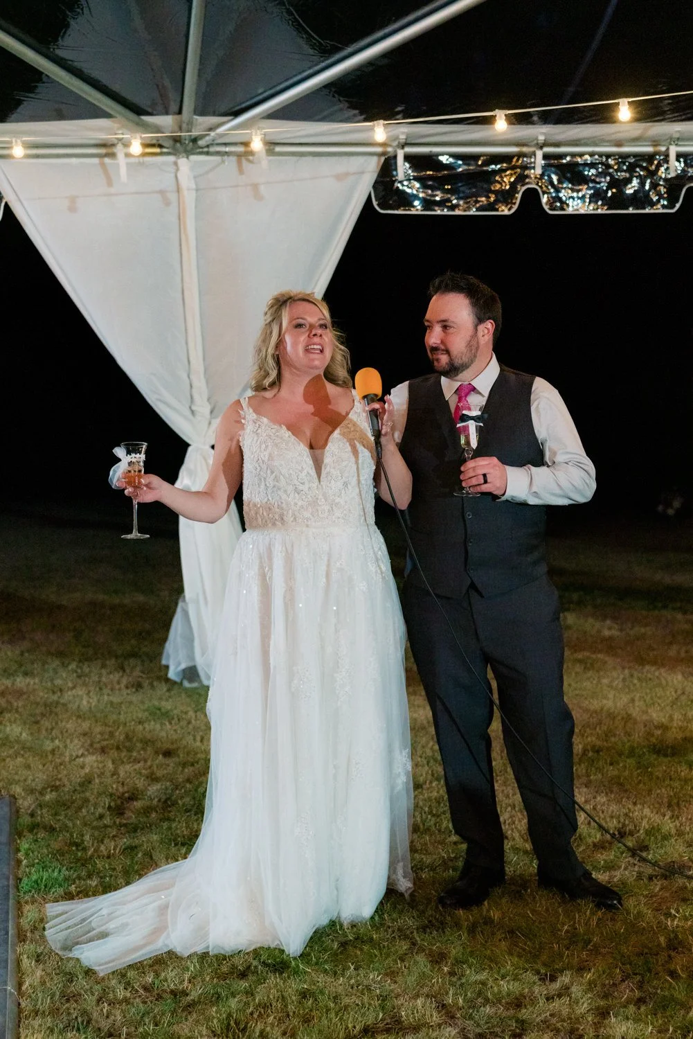 Bride giving a toast with champagne glass and microphone beside groom at an outdoor backyard wedding reception in Vancouver Washington under glowing tent lights.