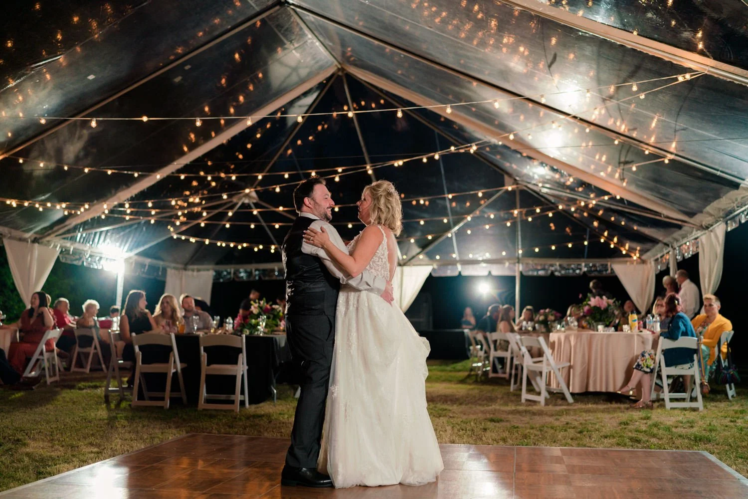 Bride and groom sharing their first dance under a clear tent with string lights at a backyard wedding in Vancouver Washington, romantic evening reception with guests seated around the dance floor.