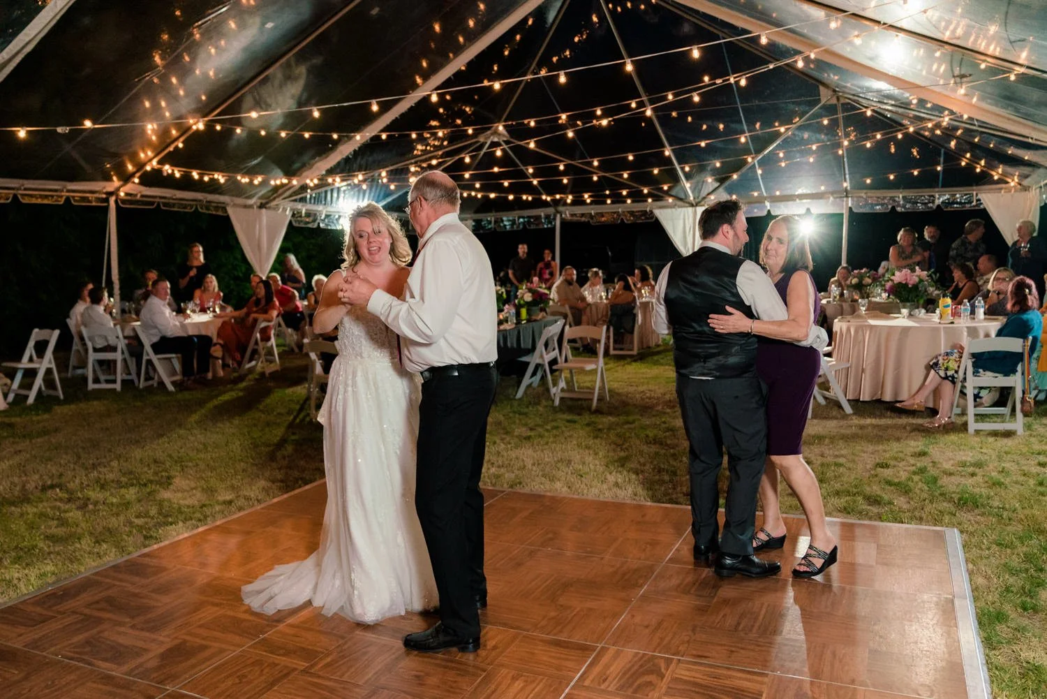 Bride and father dancing alongside another couple on a wooden dance floor at a Vancouver WA backyard wedding reception under glowing tent lights.