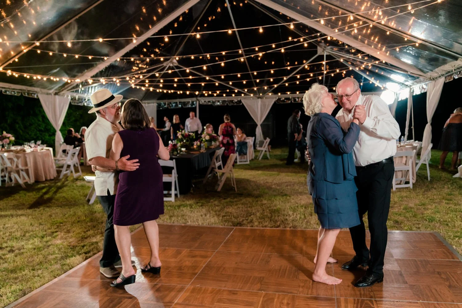 Parents of the bride slow dancing together under a clear tent with twinkle lights at a Vancouver WA backyard wedding reception surrounded by seated guests.
