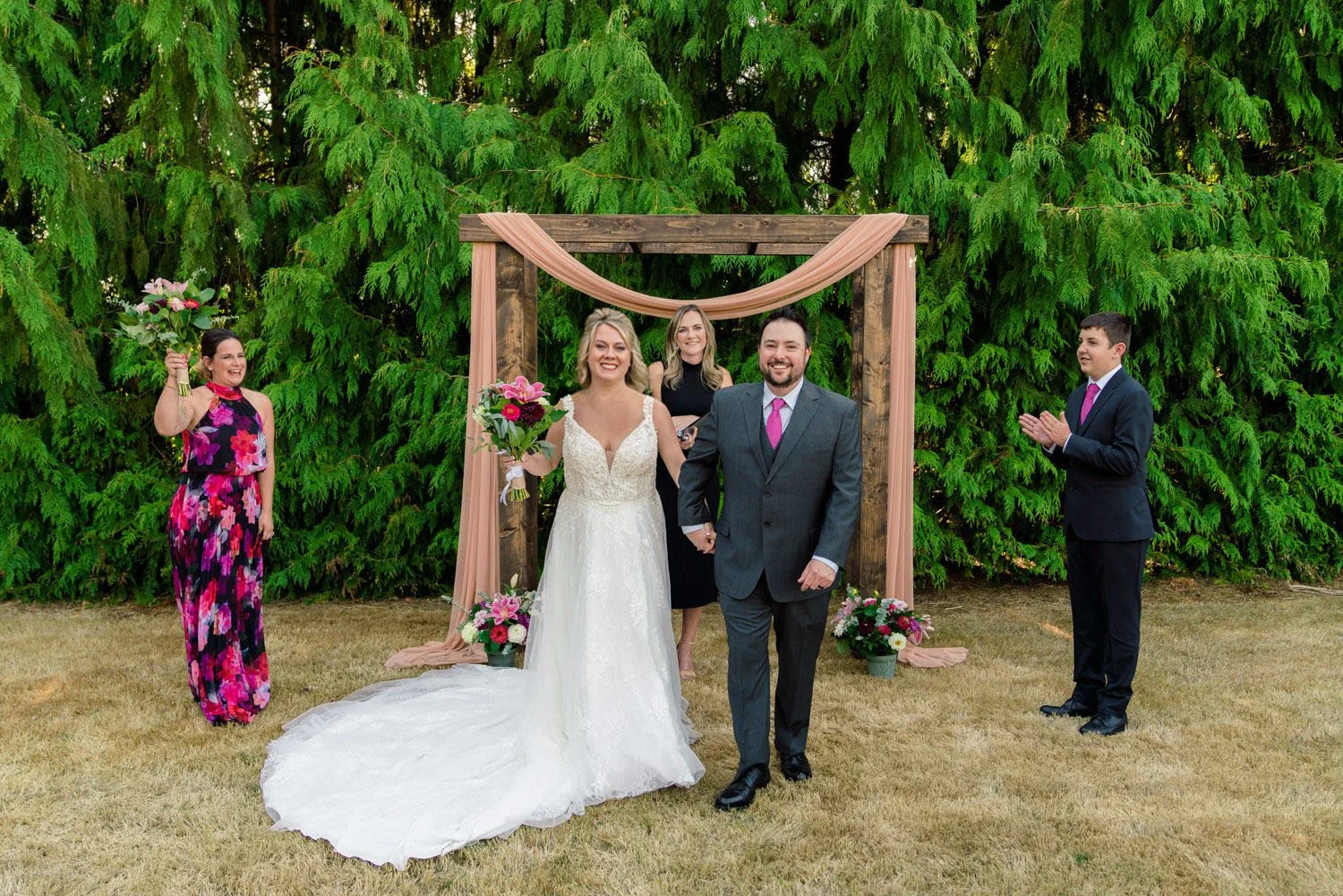 Bride and groom celebrating under wooden ceremony arch with officiant and wedding party at backyard wedding in Vancouver Washington.