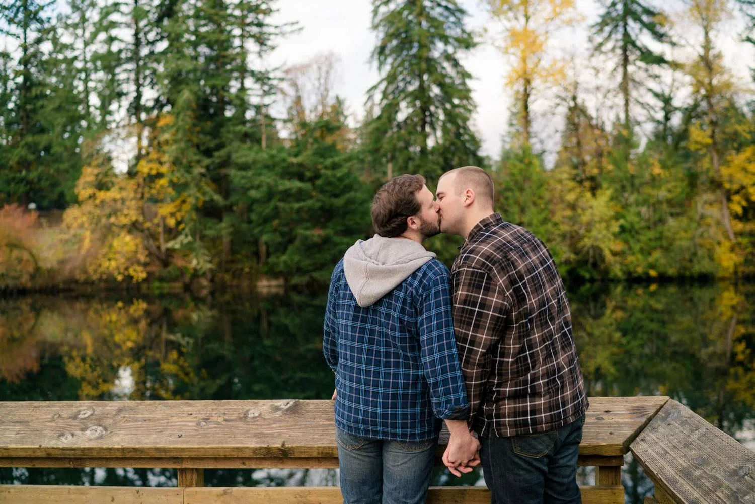 Same-sex couple engagement photos at an Oregon lake with fall trees reflected in the water.