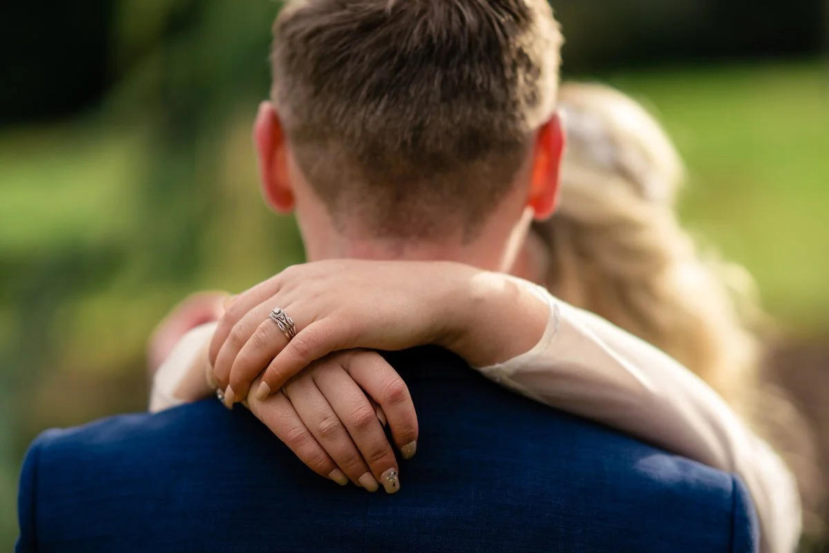 Close-up of the bride’s hands wrapped around the groom’s shoulders, showcasing her wedding ring during an intimate moment.