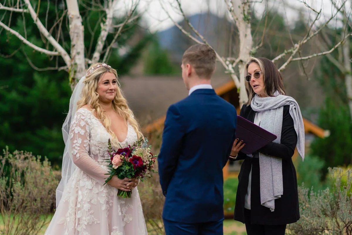 Bride holding a vibrant bouquet smiles at the groom during their outdoor wedding ceremony, with the officiant standing nearby under a serene backdrop of trees.