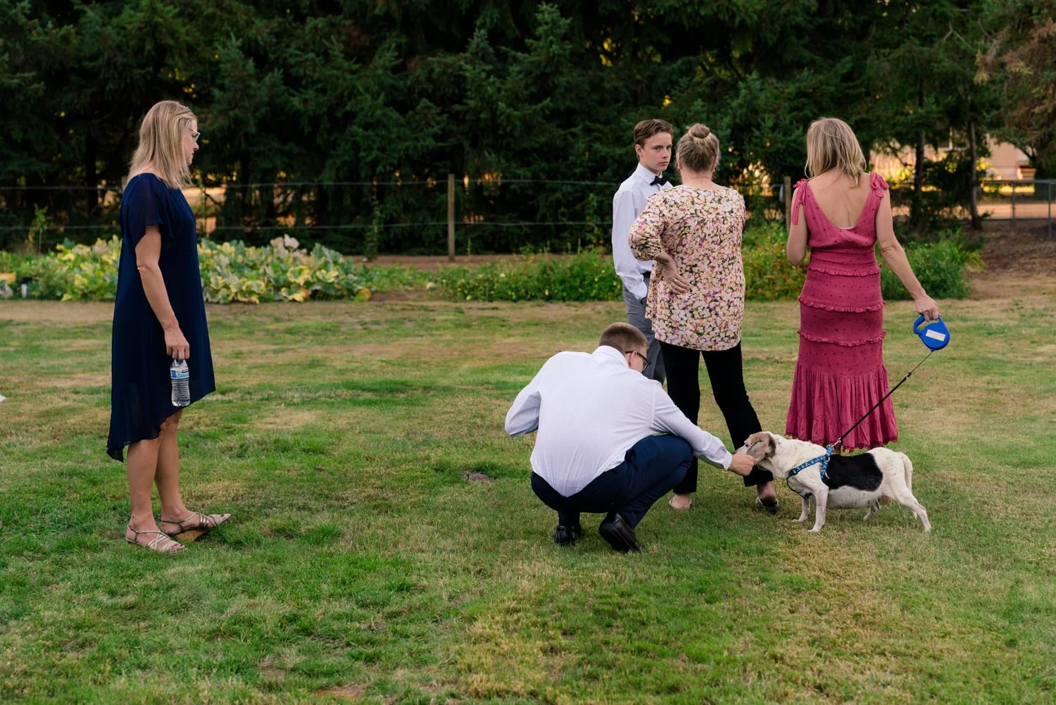Candid moment at a backyard wedding in Vancouver WA with guests chatting on the lawn while a small dog on leash gets attention before the ceremony in a relaxed outdoor setting.