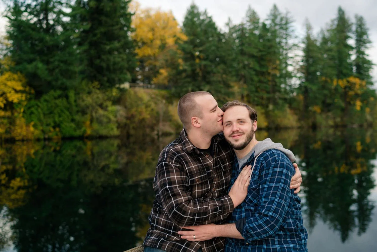 Two men sitting by a calm lake, sharing a kiss and smiling during a peaceful fall engagement session.