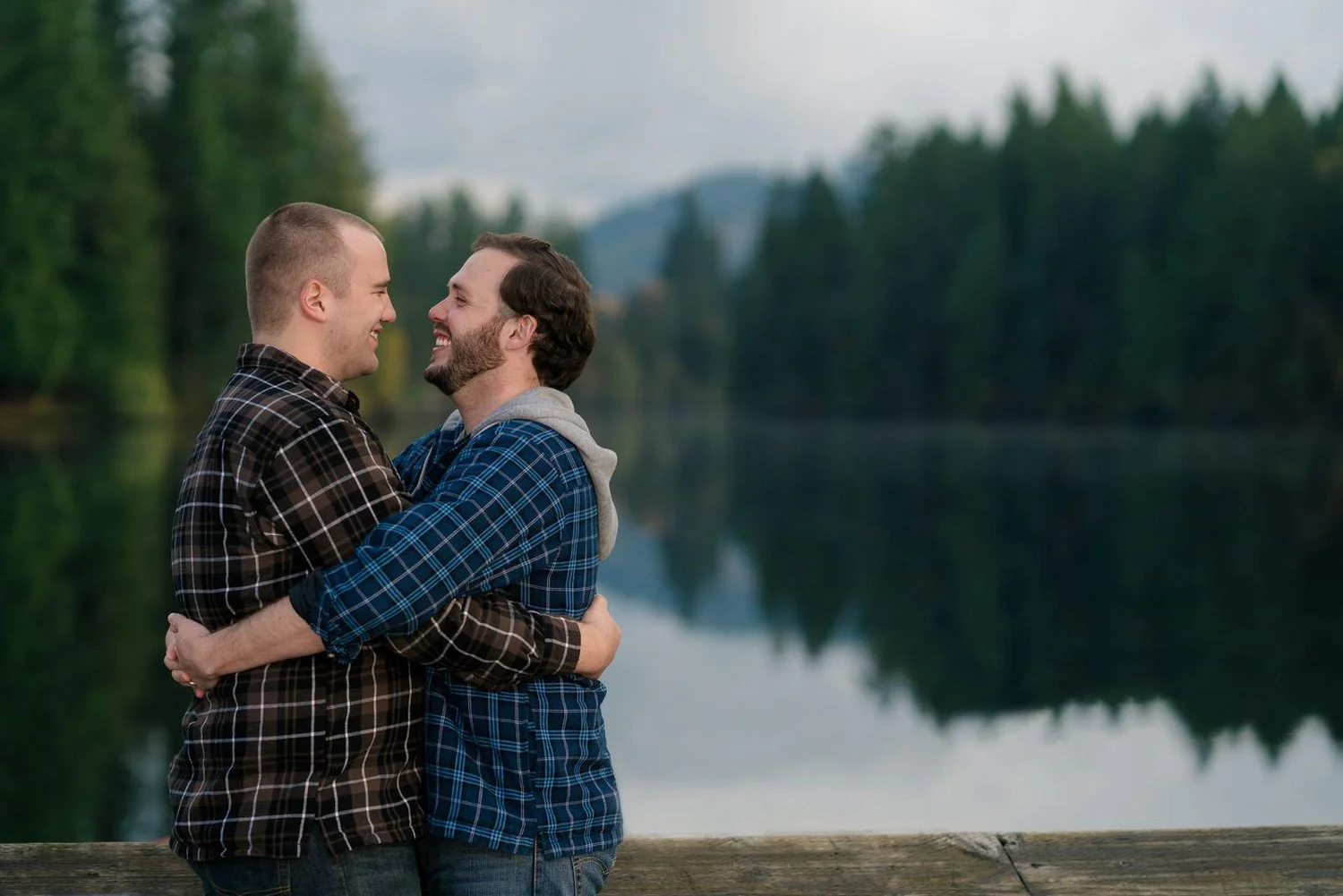 Same-sex couple engagement photos by an Oregon lake with forest reflections and mountain scenery in the background.