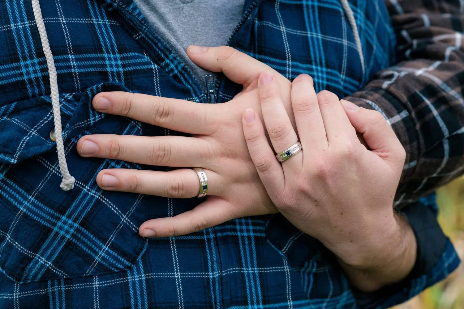 Close-up of two hands with matching rings resting over a flannel jacket during an outdoor engagement session.