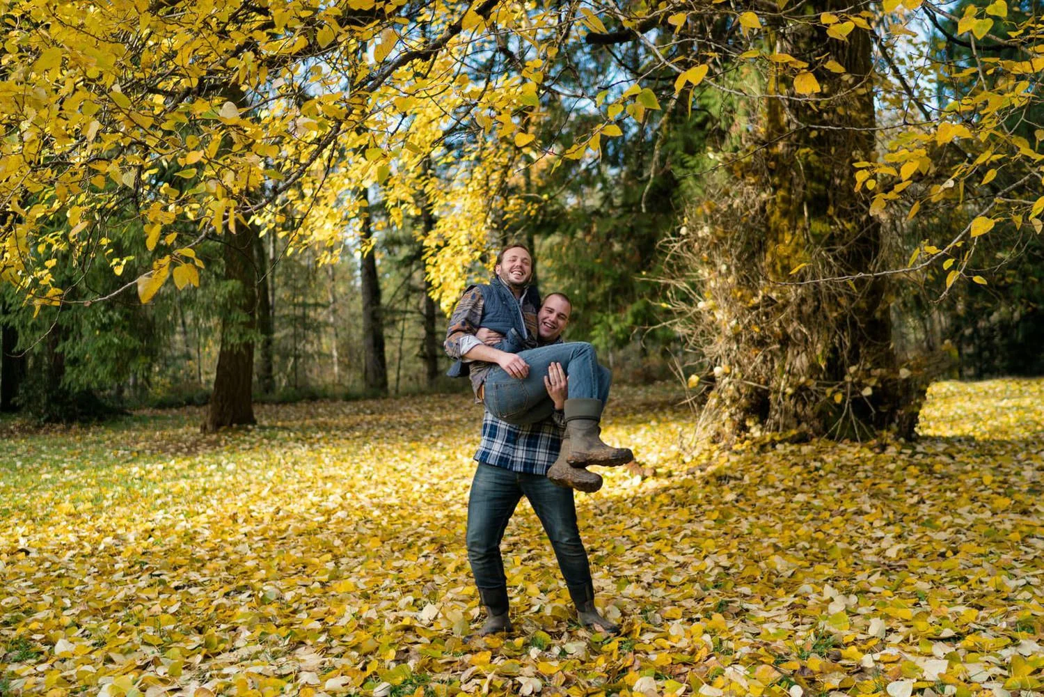 One man lifting his partner while laughing under yellow fall leaves in a forest clearing during a playful outdoor engagement session.