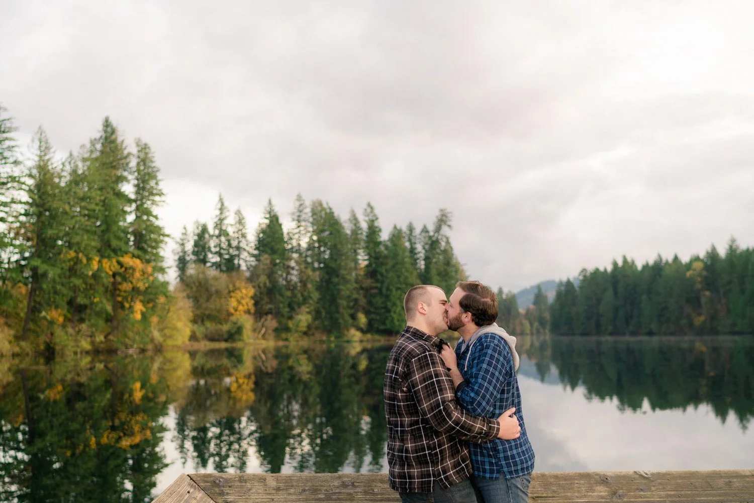 Alt text: A couple kisses during a fall engagement session at a peaceful lake in Milo McIver State Park, surrounded by evergreen trees and soft autumn colors in Oregon.