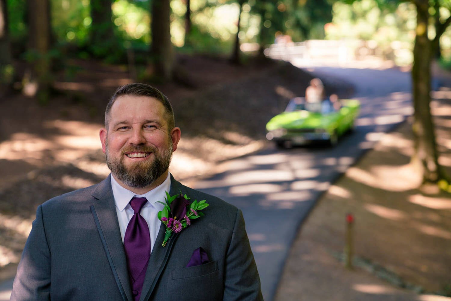 Groom smiles in a gray suit with a purple tie and boutonniere, standing on a forested path. In the blurred background, a bright green vintage convertible arrives, adding a fun and unique touch to the day.