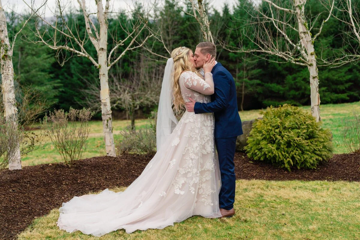 Bride and groom share a romantic kiss outdoors, surrounded by birch trees and a lush green backdrop on their wedding day.