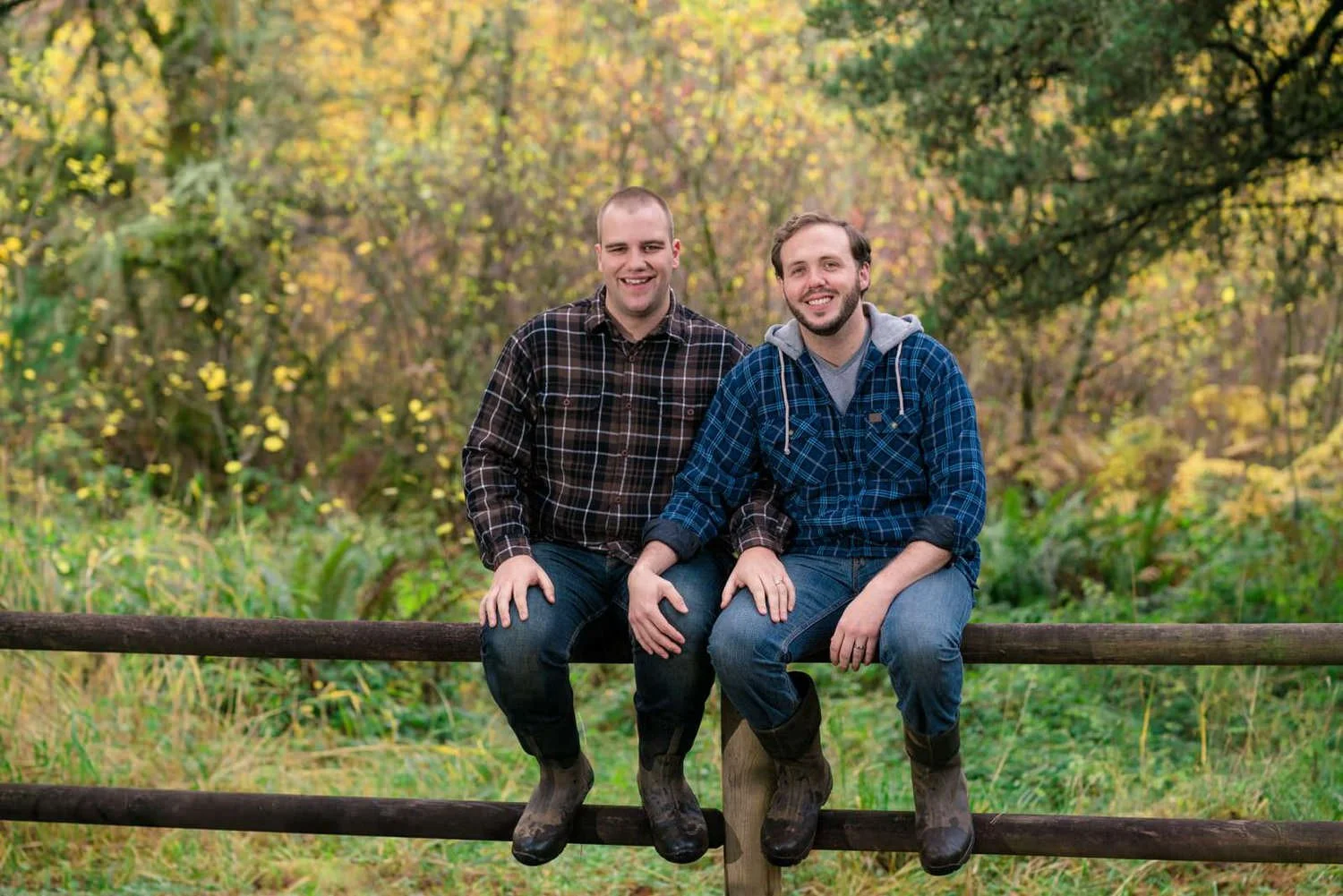 Two men sitting on a wooden fence and smiling at the camera in a green forest during a relaxed fall engagement session.