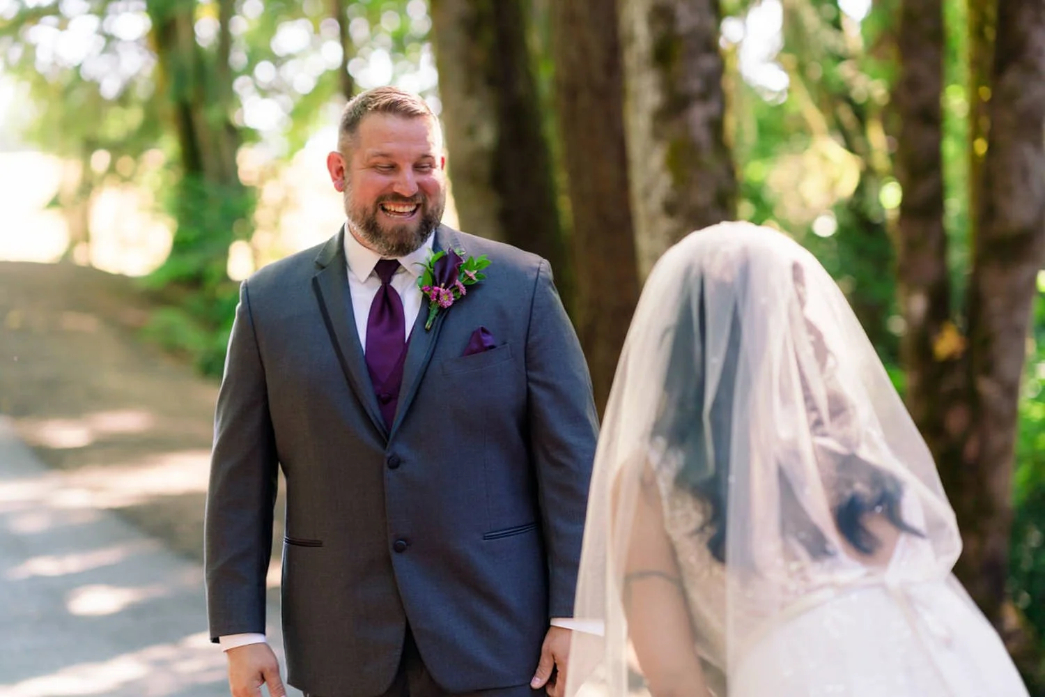 Groom smiles brightly as he sees his bride for the first time during their emotional first look. The bride, wearing a veil and sparkly gown, stands just in front of him under the forest canopy.