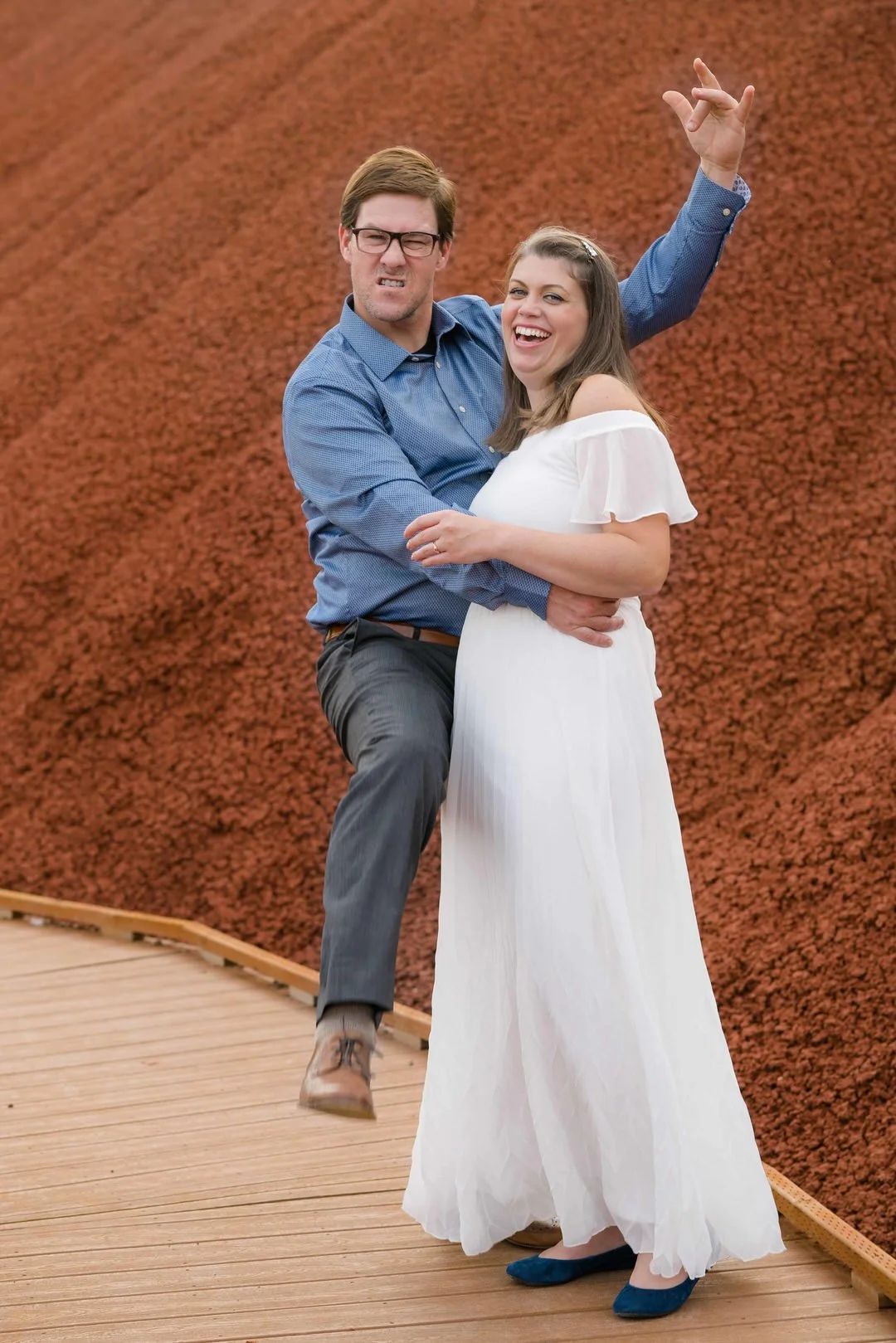 Playful couple posing on a wooden boardwalk at the Painted Hills, with the man making a funny expression and lifting his leg while the woman laughs in a flowing white dress.
