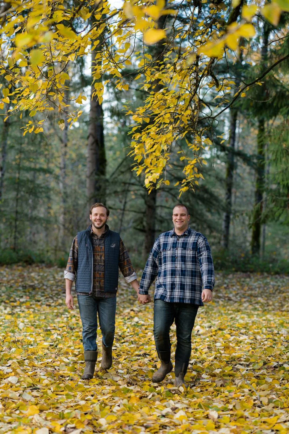 Two men holding hands and walking through a forest covered in golden fall leaves during an outdoor engagement session.