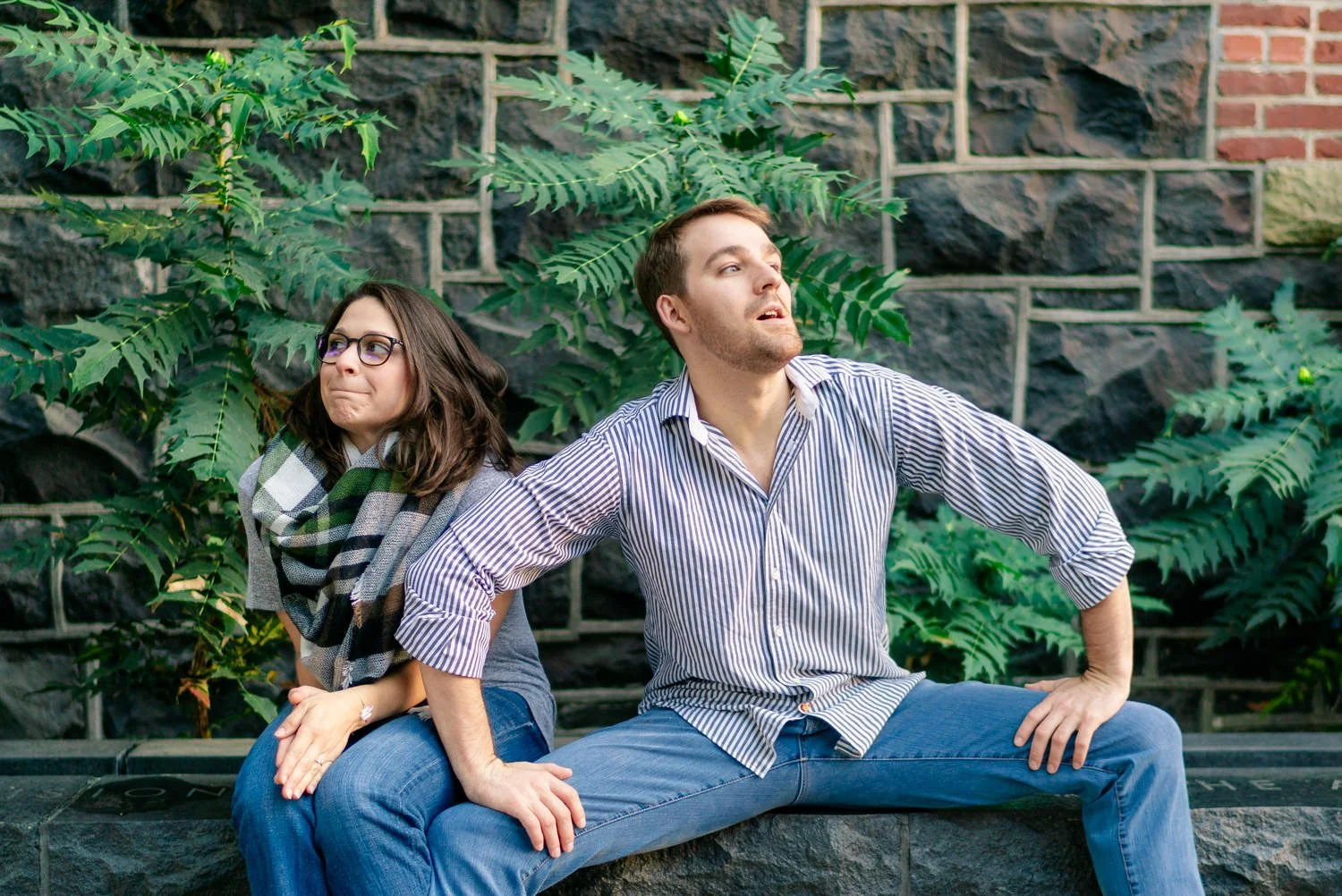 Couple making playful faces while sitting on a stone ledge in front of a textured wall and greenery during their fun and quirky Portland engagement session.
