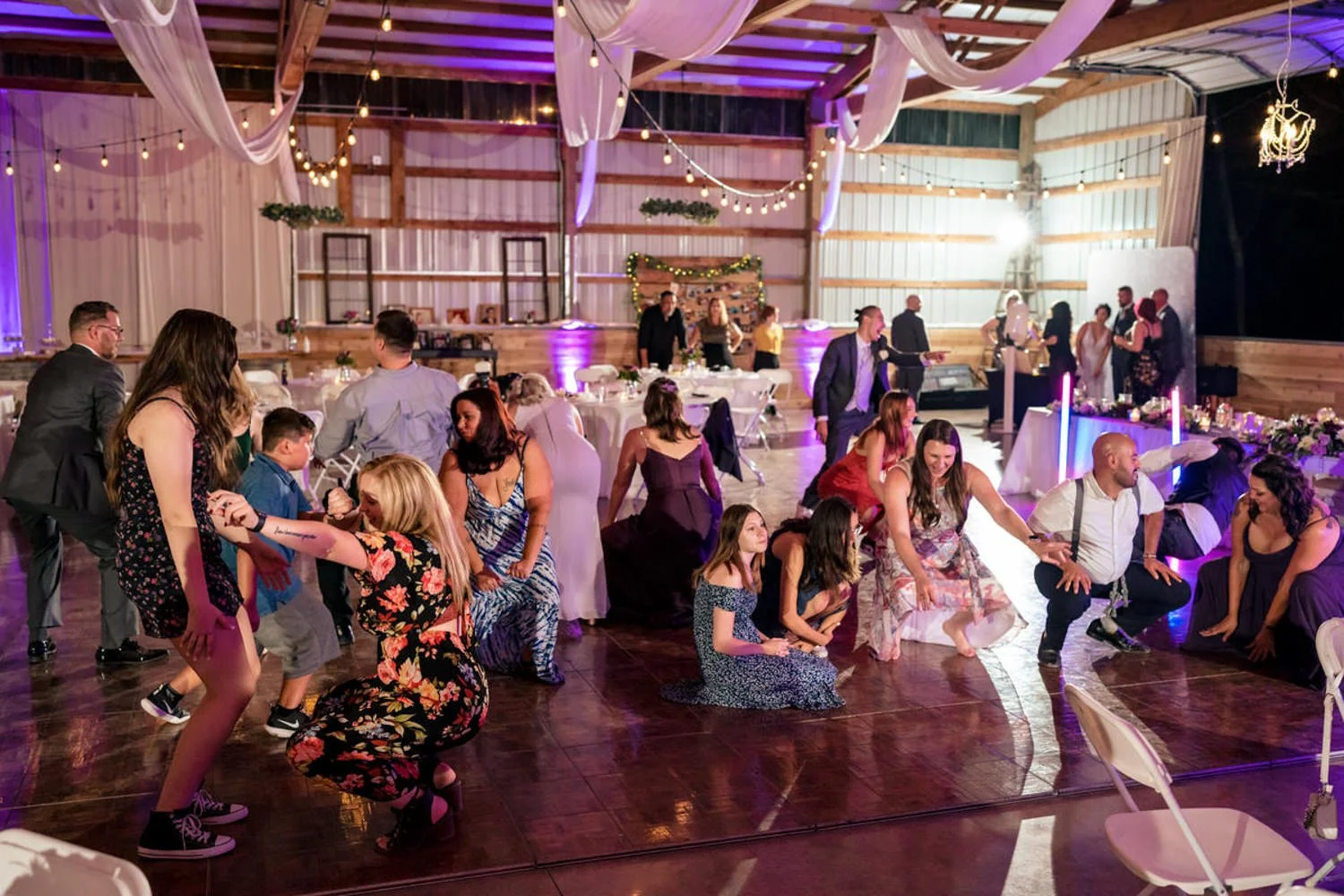 Guests crouch and dance in sync on the barn wedding reception floor during a fun group dance moment. Colorful lights, string lights, and draped fabric create a festive and lively atmosphere.