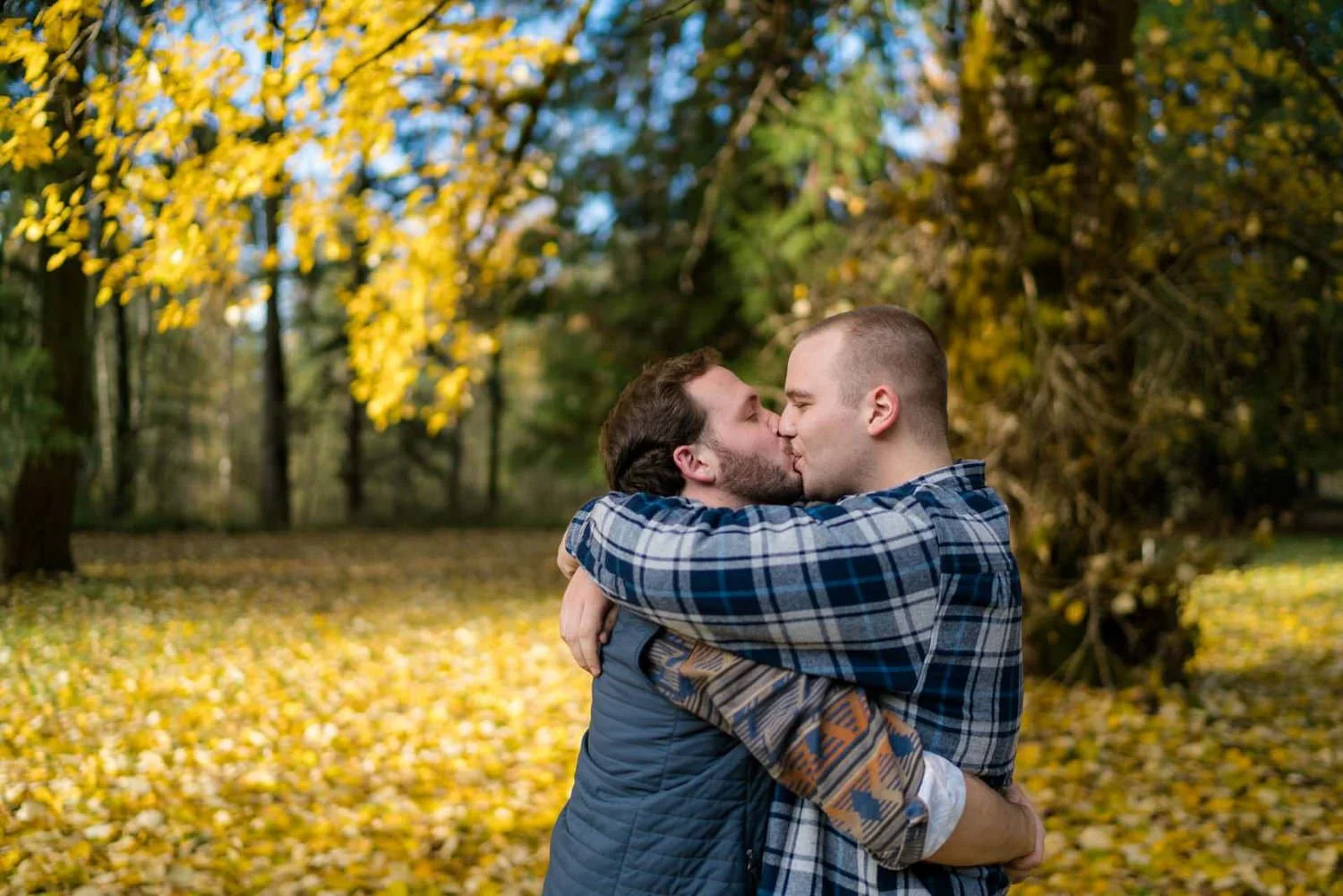 Two men embracing and kissing in a forest surrounded by golden autumn leaves during an outdoor fall engagement session.