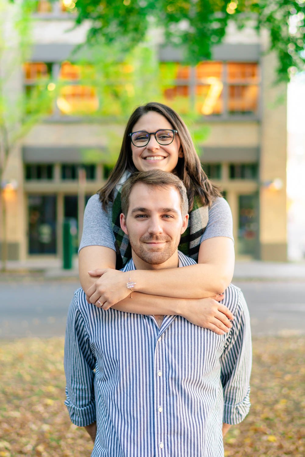 Couple smiling during their Portland engagement session, with one partner giving the other a sweet hug from behind in a cozy, tree-lined urban setting.
