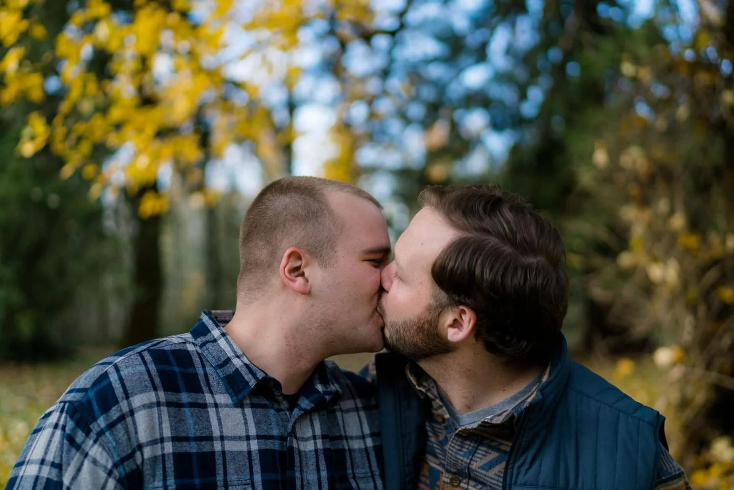 Two men sharing a kiss on a wooded trail with golden fall leaves, captured during an outdoor engagement session in autumn.