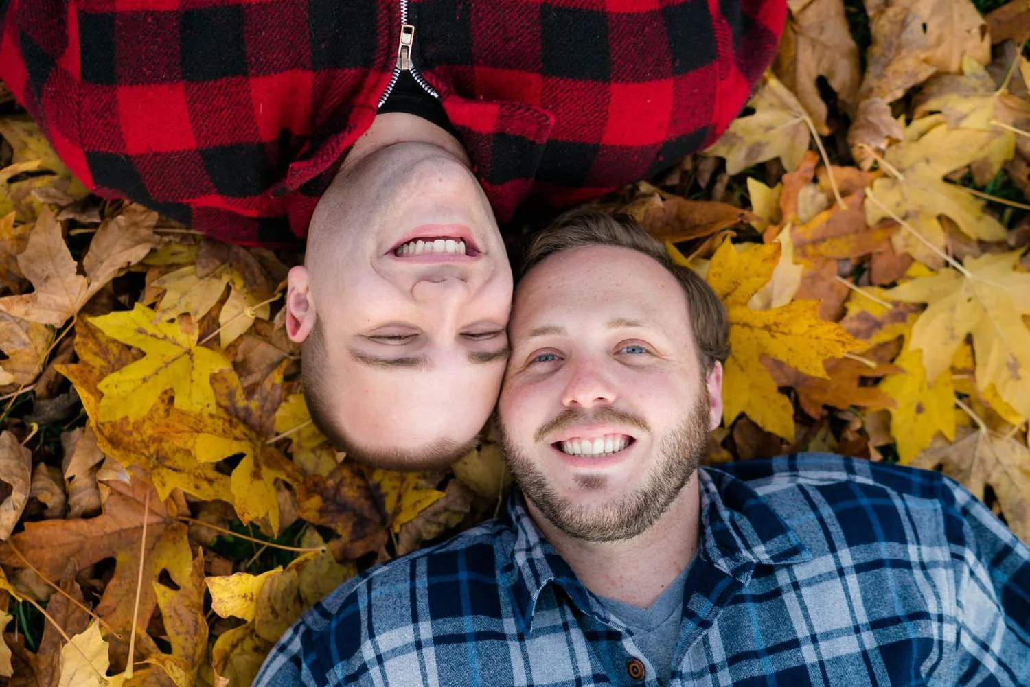 Two men lying in fallen autumn leaves and smiling at the camera during a cozy outdoor engagement session.