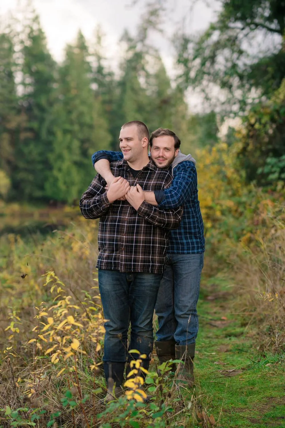 One man hugging his partner from behind while standing on a forest trail during a relaxed fall engagement session.