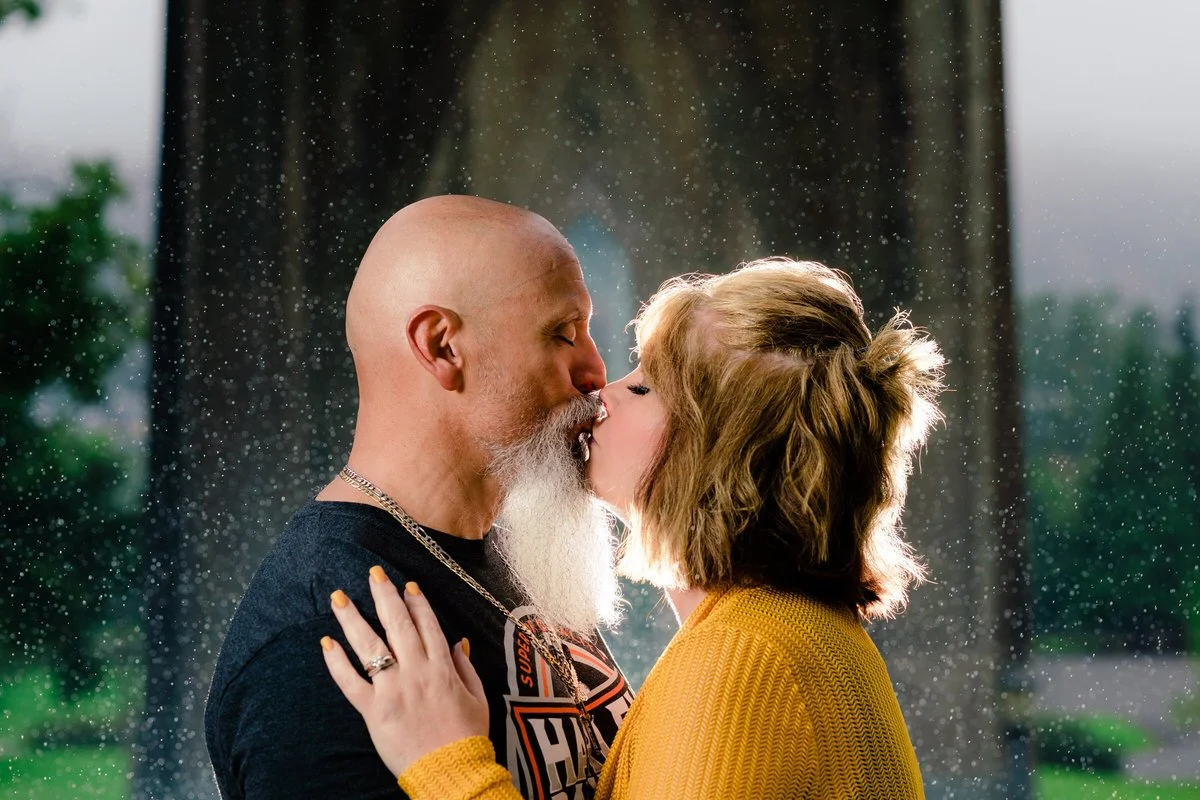 A romantic kiss under the arches of Cathedral Park: a bald man with a white beard kisses a blonde woman in a yellow sweater, with shimmering raindrops in the background.