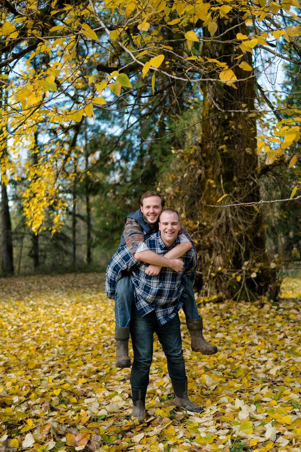 One man giving his partner a piggyback ride in a forest filled with golden fall leaves during a fun outdoor engagement session.