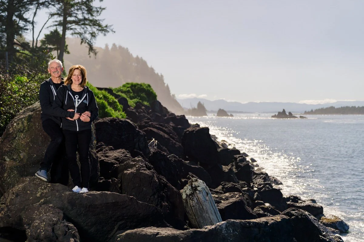 A smiling couple wearing matching black hoodies stands on rocky terrain near a scenic coastline, with sunlight glinting off the water.