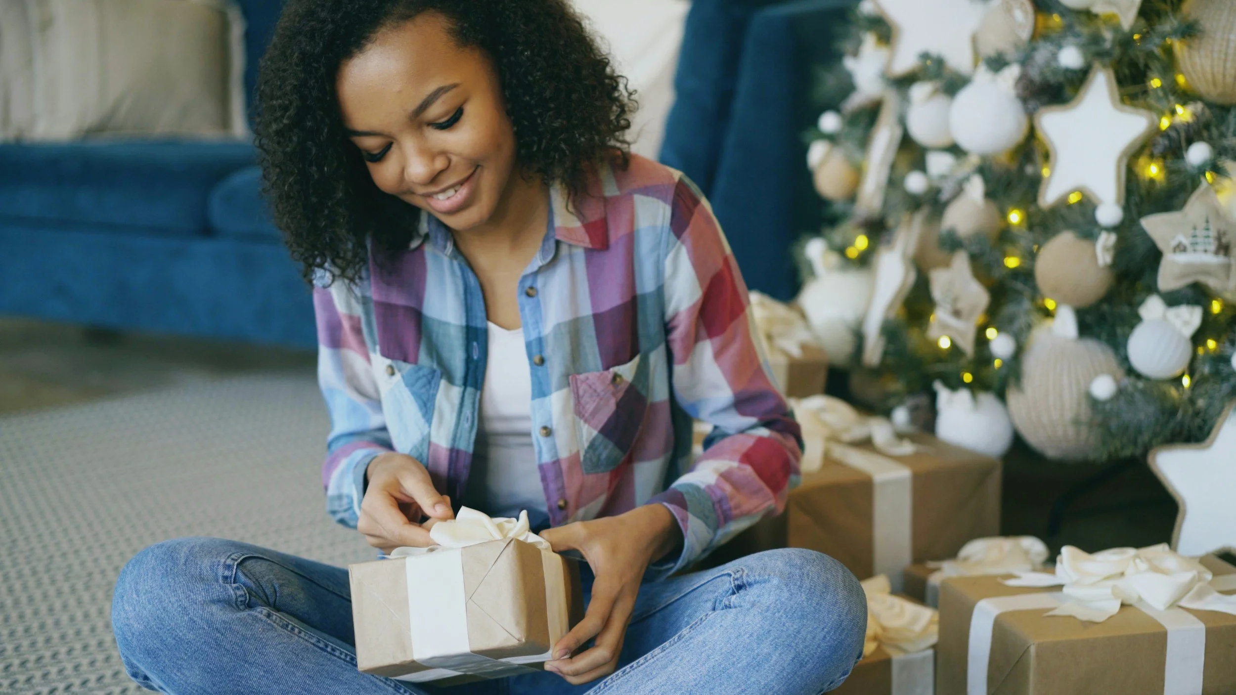 A woman sitting cross-legged on the floor, wrapping a gift with a beige ribbon, near a decorated Christmas tree with white ornaments and gift boxes around her.