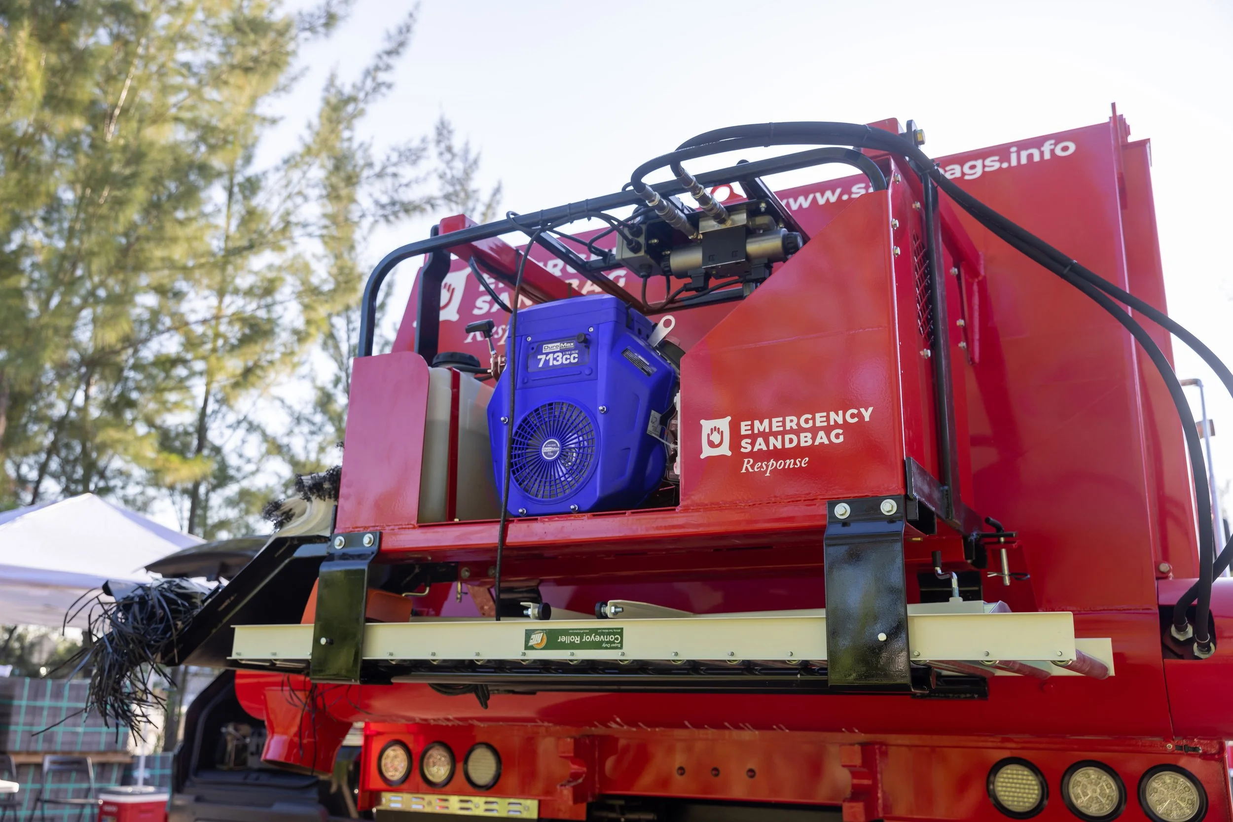 Fire truck with an emergency sandbag response system, featuring a blue engine, mounted on the back, with trees and tents in the background.