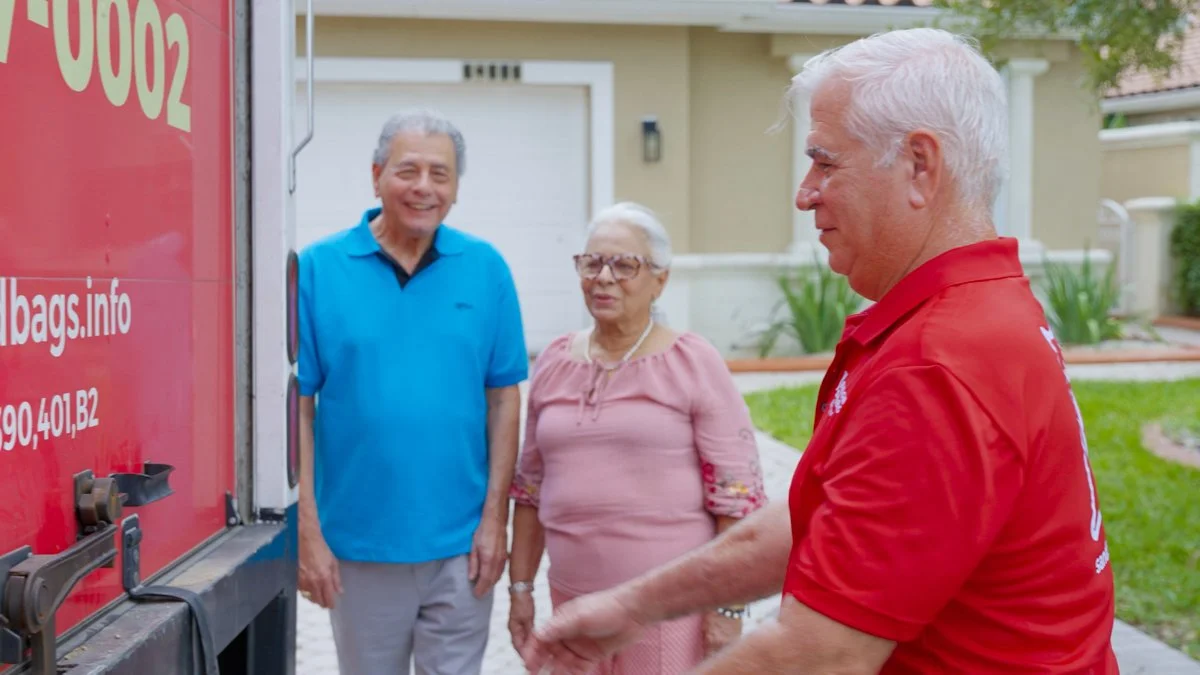 A food delivery person in a red shirt stands in front of two elderly people, a man in a blue polo shirt and a woman in a pink blouse, who are smiling and looking at him outside a residential house with a green lawn.