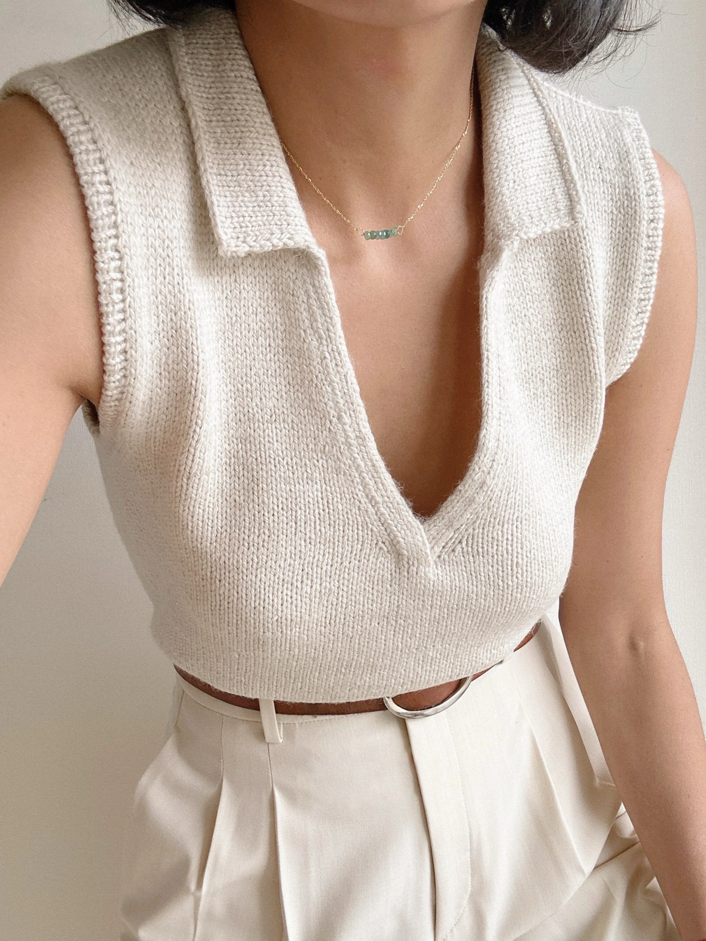A woman wearing a knit sleeveless cream top, white high-waisted pants, a delicate gold necklace, and a brown leather belt.