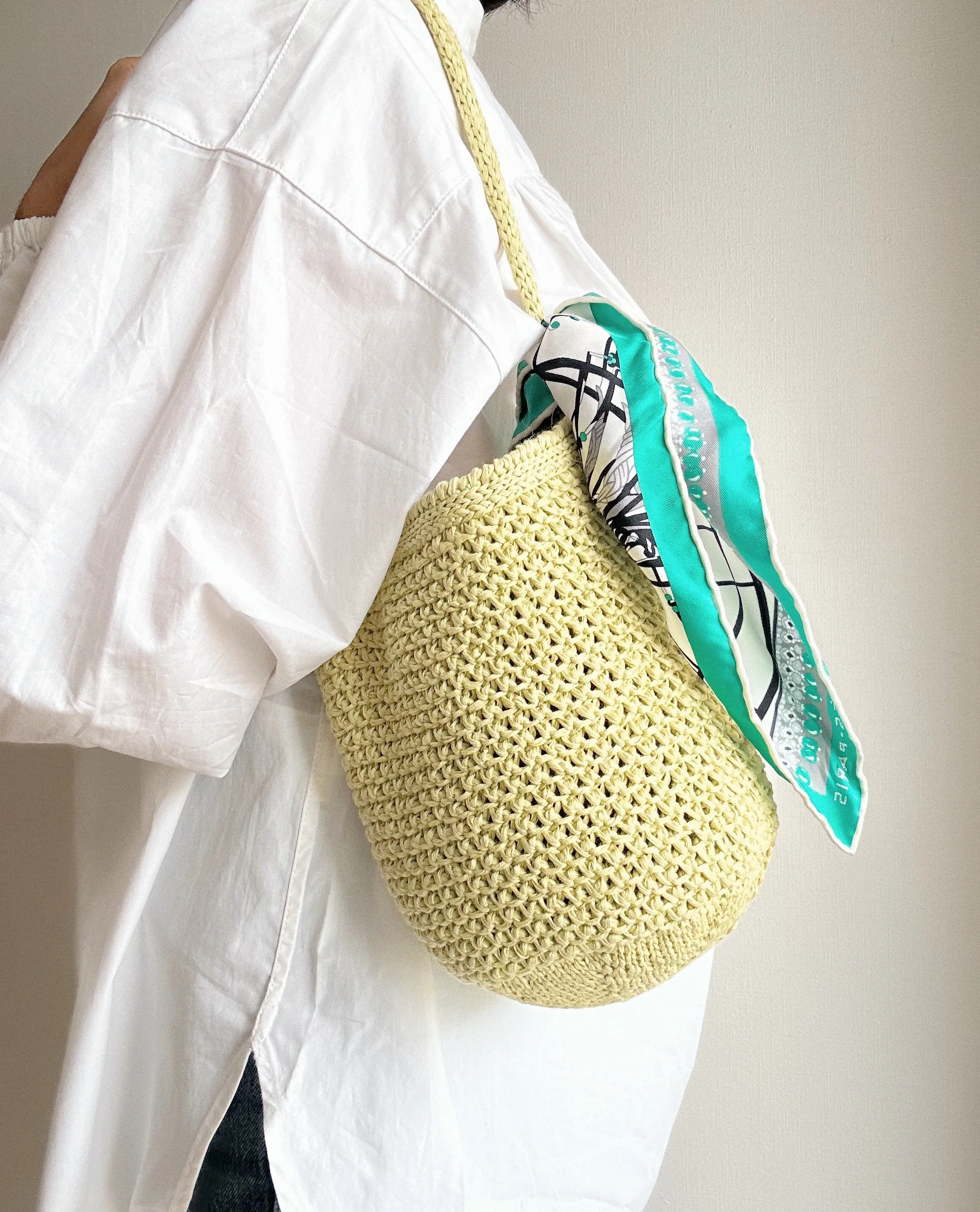 A person wearing a white shirt with a cut-out shoulder, carrying a large yellow crochet bag with a colorful scarf tied to its handle.