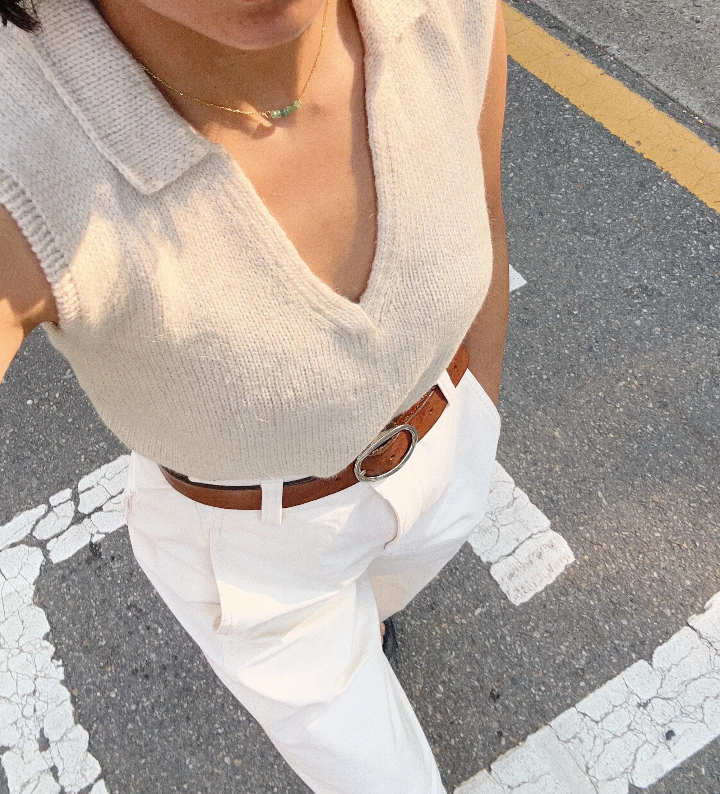 Top-down photo of person standing on street in white pants, brown belt, beige sleeveless knit top, and a necklace, with a cracked parking lot surface and yellow lines.