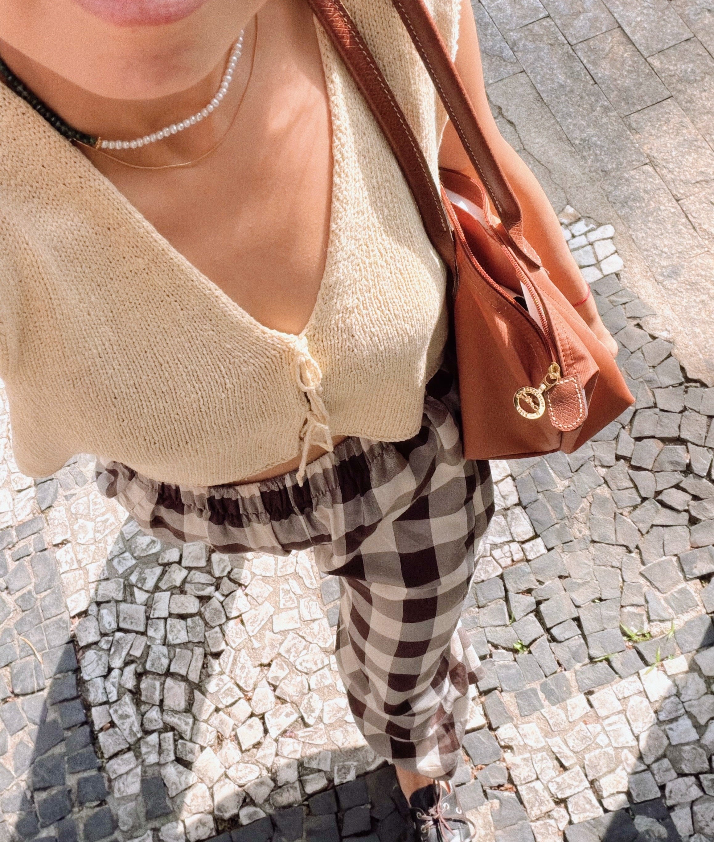 A top-down view of a person standing on cobblestone pavement, wearing a beige knit cardigan, black and white checkered pants, gray shoes, a pearl necklace, and a brown handbag.