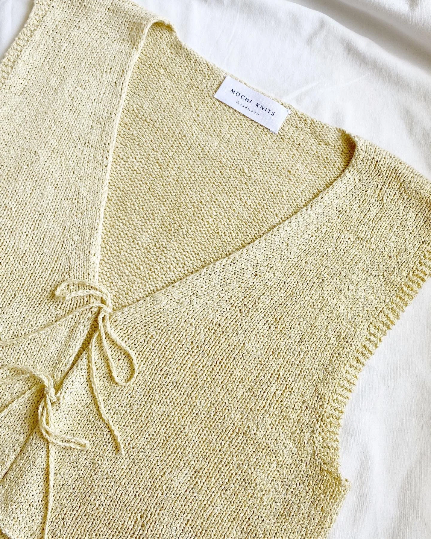 Light yellow knitted garment with a tag that reads 'Mochi Knits Handmade' and a drawstring tie.