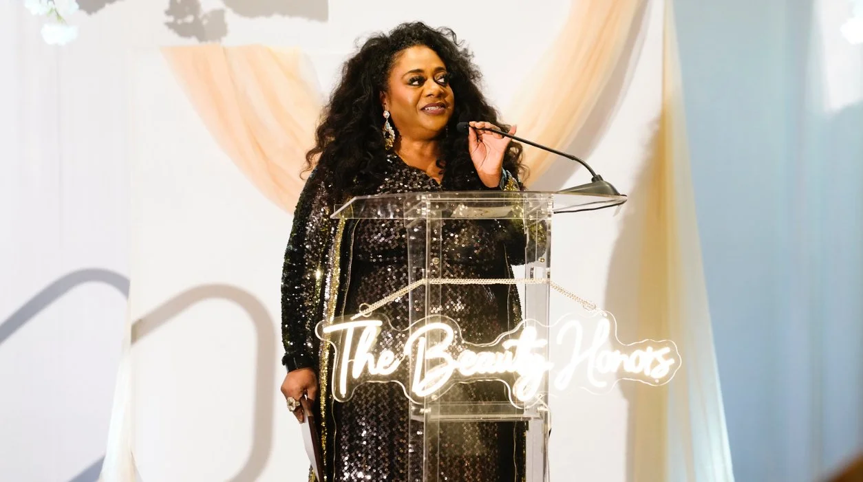 Woman in red off-the-shoulder dress speaking at a podium with red drapes and white background.