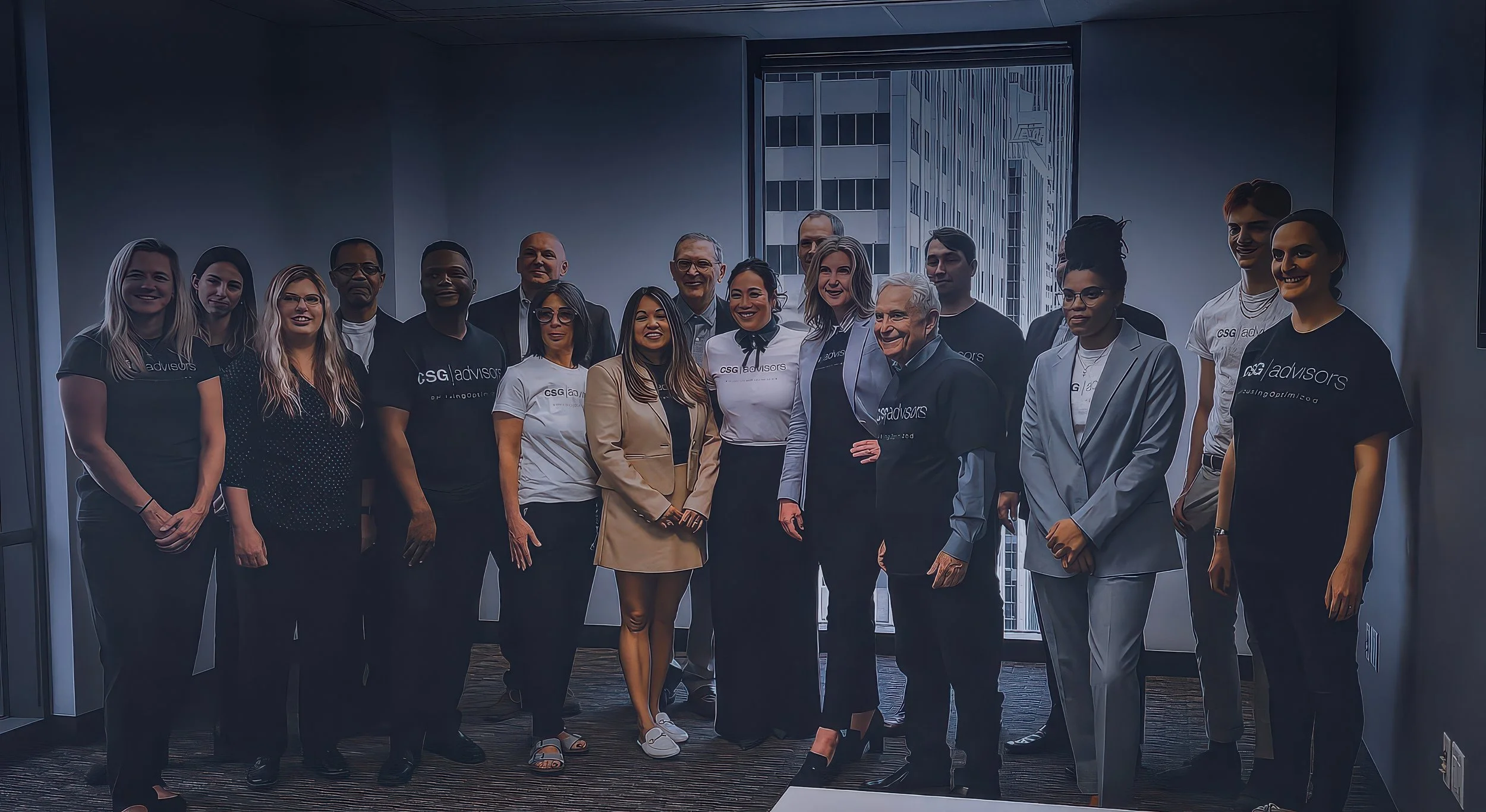 Group of diverse professionals, some wearing CSG Advisors t-shirts, standing together indoors in front of large windows with city skyscrapers outside.