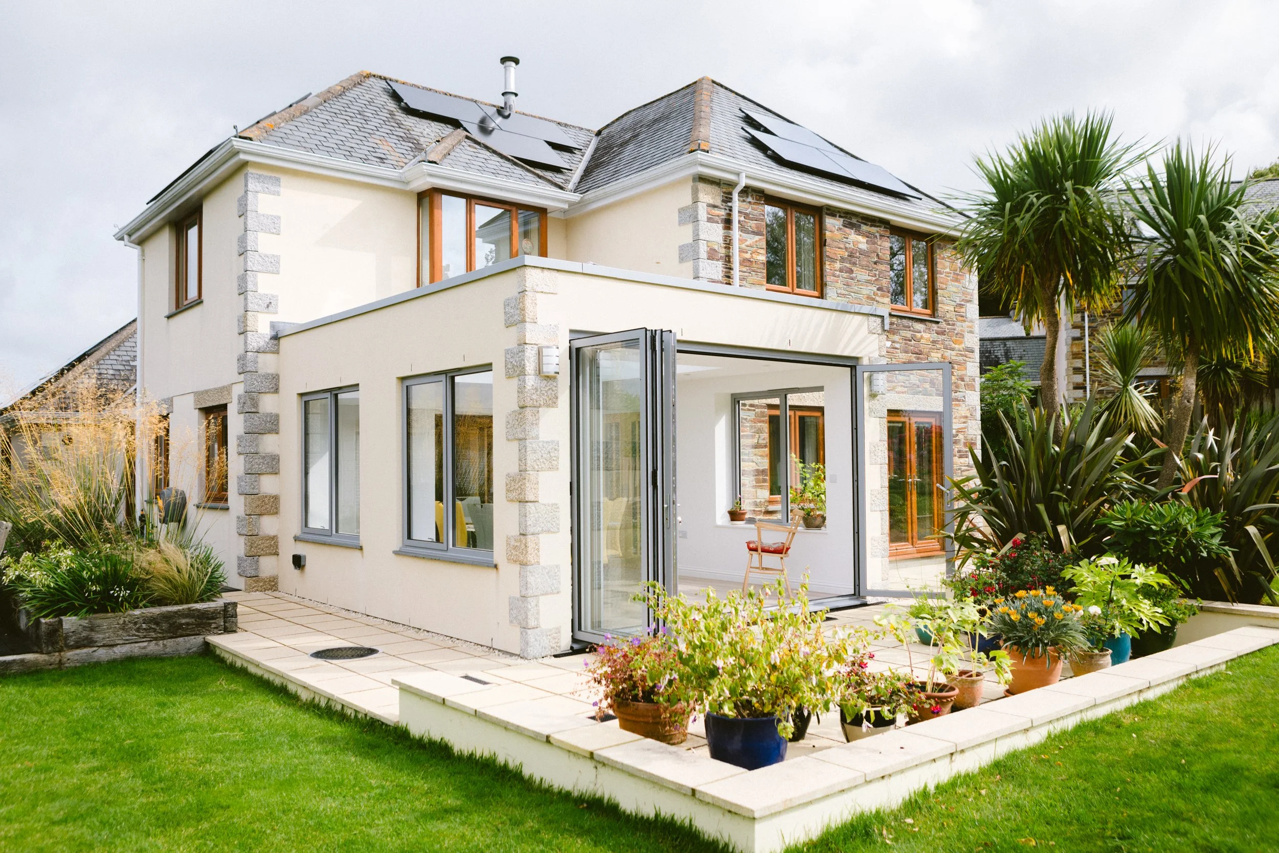 A modern two-story house with solar panels on the roof, surrounding garden with various potted plants, and a partially open glass extension in the backyard.